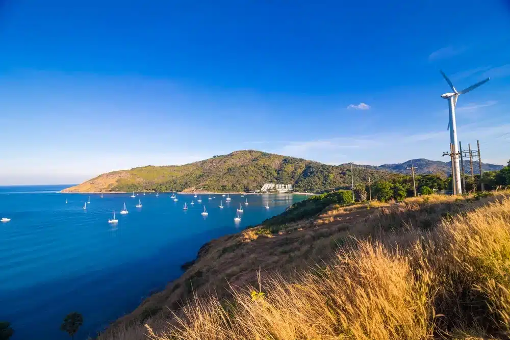 A scenic coastal view at Laem Phromthep showcases a bright blue sky over a tranquil bay with numerous sailboats anchored in the calm, blue water. A hill covered in dry grass occupies the foreground, while a modern wind turbine stands to the right. Lush green hills and a few buildings are visible in the background.