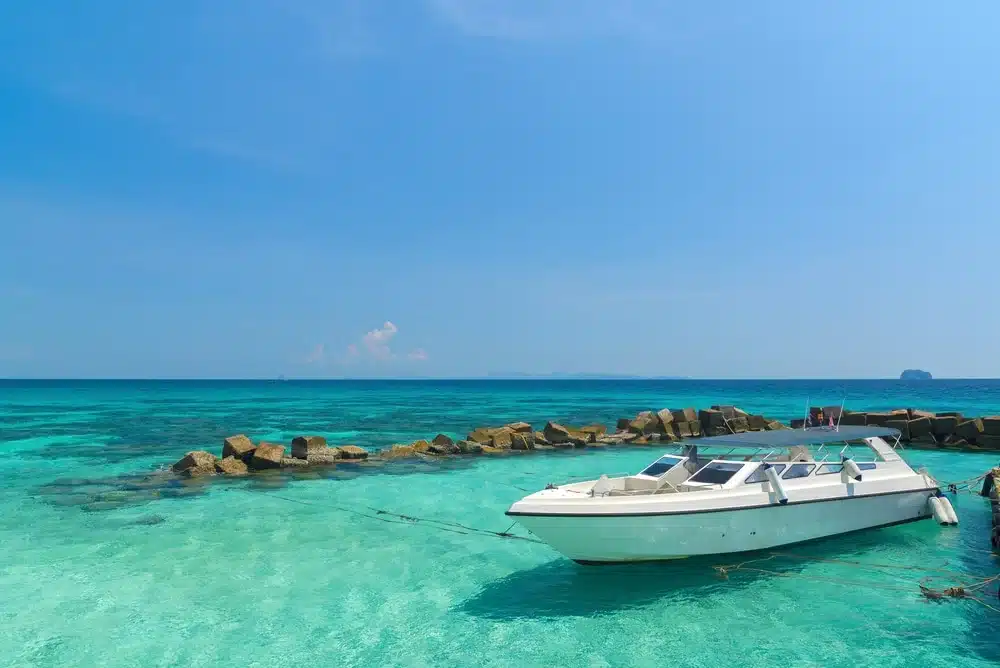 A white boat floats on crystal-clear turquoise water near Patong Beach. The boat is anchored and surrounded by scattered rocks, forming a small protective barrier. In the background, the horizon merges with a clear blue sky, creating a serene ocean scene—a perfect escape from the lively Patong nightlife.