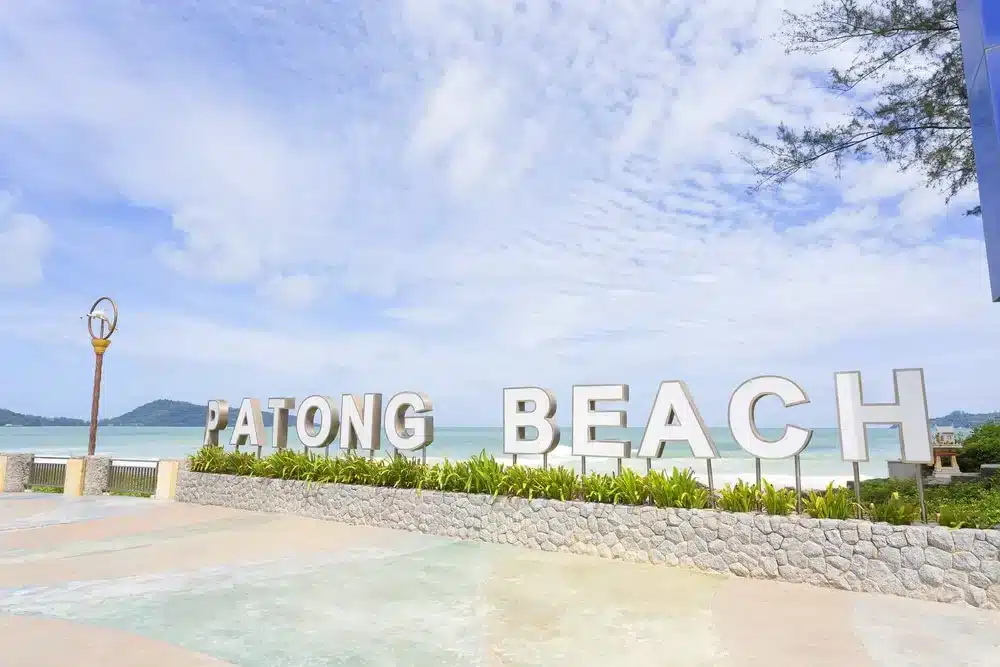 Large white letters spell out "Patong Beach" on a stone wall along a sunny, sandy coast. The ocean is calm and turquoise beneath a partly cloudy sky. Greenery grows at the base of the wall. Trees and mountains, reminiscent of those near Wat Chalong Phuket, are visible in the distance, and a decorative lamppost stands to the left.