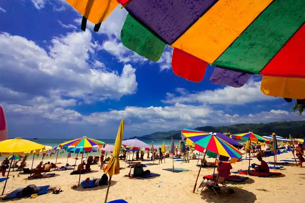 A bustling beach scene under a bright blue sky with scattered clouds unfolds at one of the best places to visit in Patong. Colorful umbrellas in shades of green, yellow, red, and purple provide shade to numerous people lounging on the sandy shore. The lively beachgoers enjoy gentle waves visible in the background.