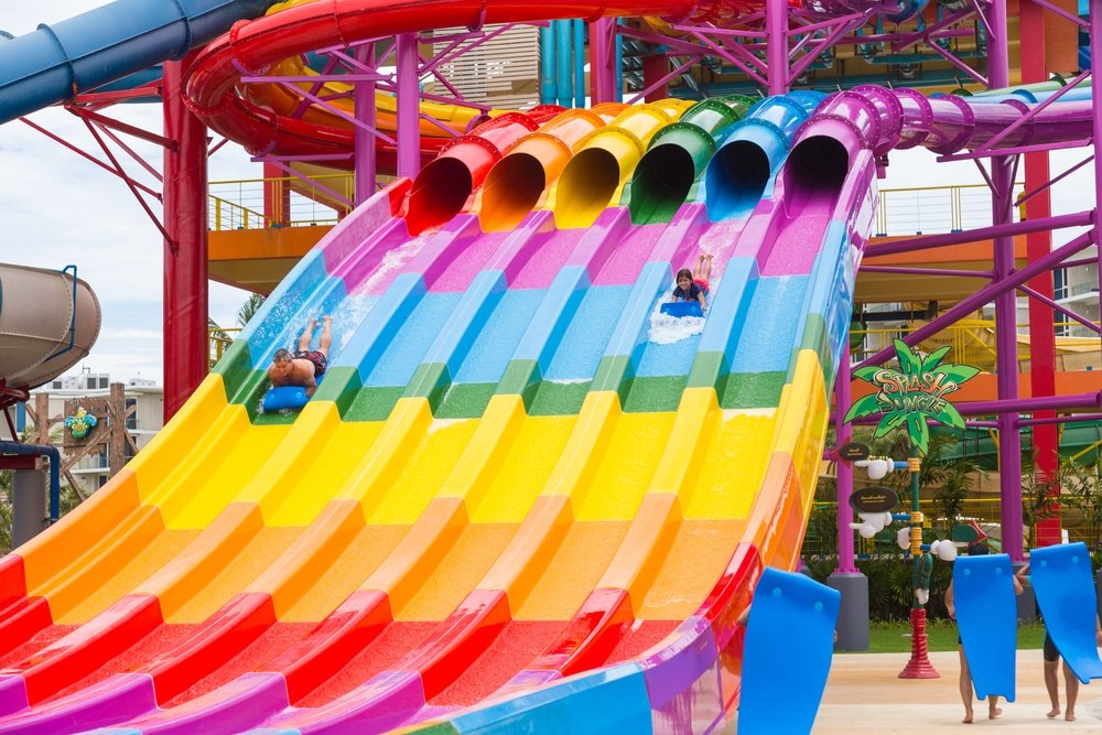 A vibrant, multi-lane rainbow water slide with several people sliding down is among the top Phuket attractions. The slide features bright sections in red, orange, yellow, green, blue, and purple. The energetic atmosphere is enhanced by colorful tubes overhead and lively park decorations in the background.