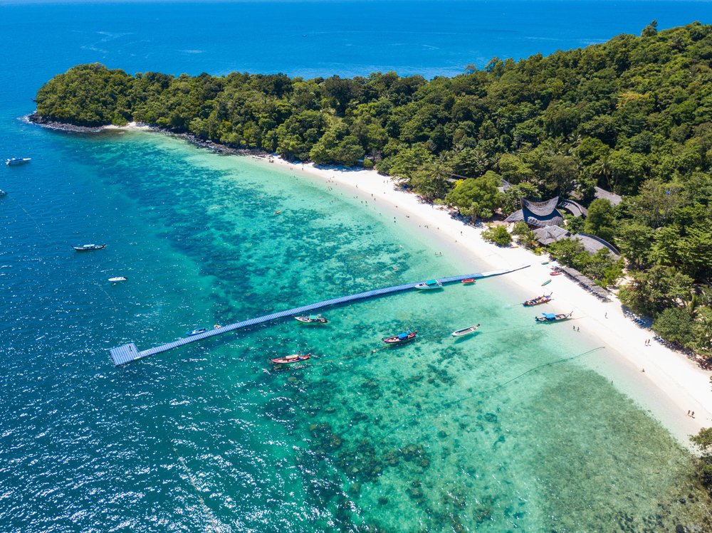 Aerial view of a pristine beach in Phuket with turquoise waters and lush greenery. A floating pier extends into the water, where several boats are moored. People are seen enjoying the beach, while dense forest surrounds the shoreline and extends into the background. Sunny weather and clear skies enhance this Phuket travel guide scene.