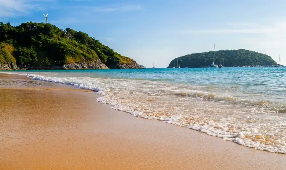 A serene beach scene with gentle waves lapping against the sandy shore. Two green, tree-covered hills are visible in the background under a partly cloudy sky. A wind turbine is on the left hill, and several sailboats are anchored near the right hill in the azure water—much like Patong Beach attractions.