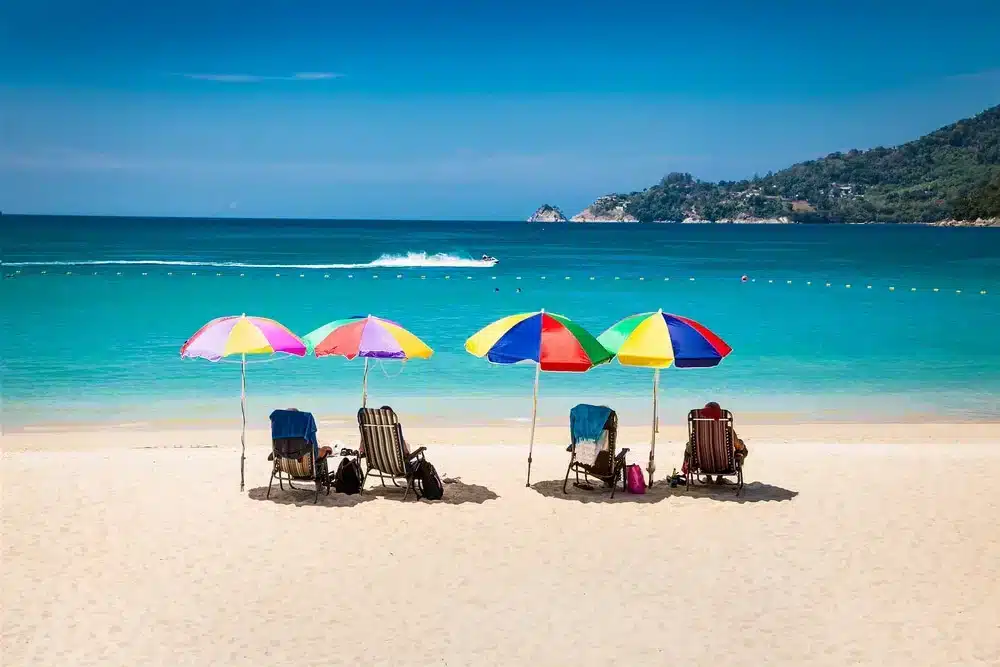 Four colorful beach umbrellas provide shade for lounge chairs on a sandy beach. The blue ocean water in the background features a speedboat creating waves. Distant hills with lush greenery, reminiscent of Wat Chalong Phuket, are visible under a clear, sunny sky. Floating buoys mark a swimming area.