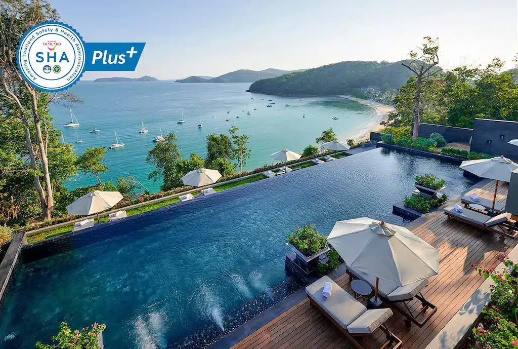 A luxurious infinity pool with adjacent deck chairs and umbrellas overlooks a serene ocean at one of the best hotels in Phuket near the beach. Sailboats are visible on the clear blue water, and green hills border the sandy beach. The image also includes a "SHA Plus+" certification logo in the top left corner.