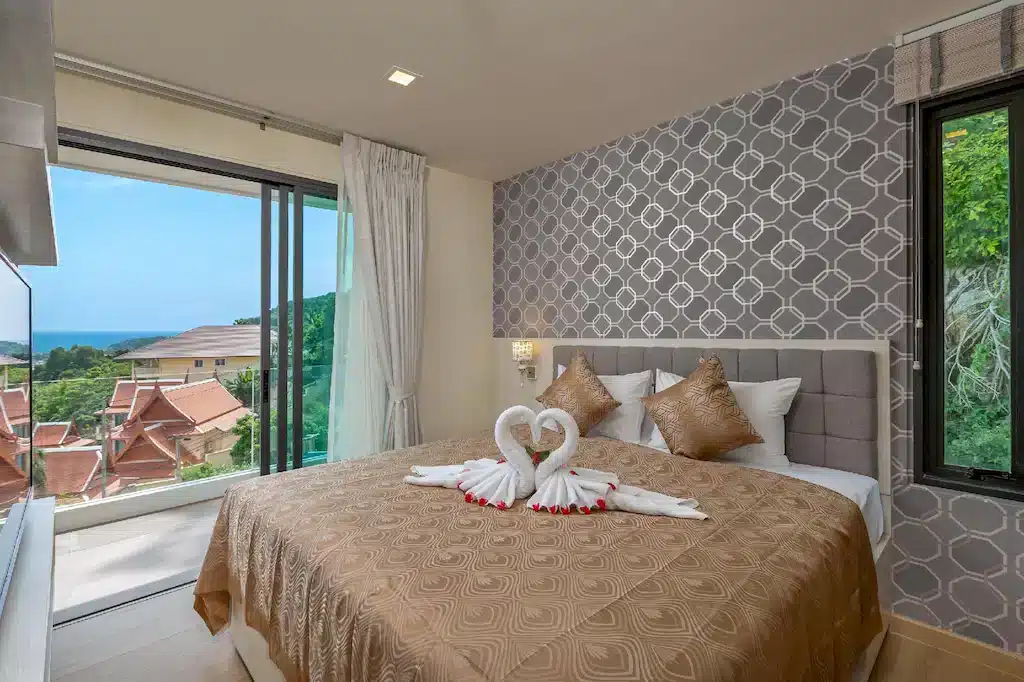 A bedroom with a large window offering a view of a coastal area with traditional-style buildings and the distant silhouette of Big Buddha Phuket. The bed has a gold-patterned bedspread and four matching pillows. Two towel swans form a heart on the bed, surrounded by red rose petals. The walls are decorated with grey geometric-patterned wallpaper.