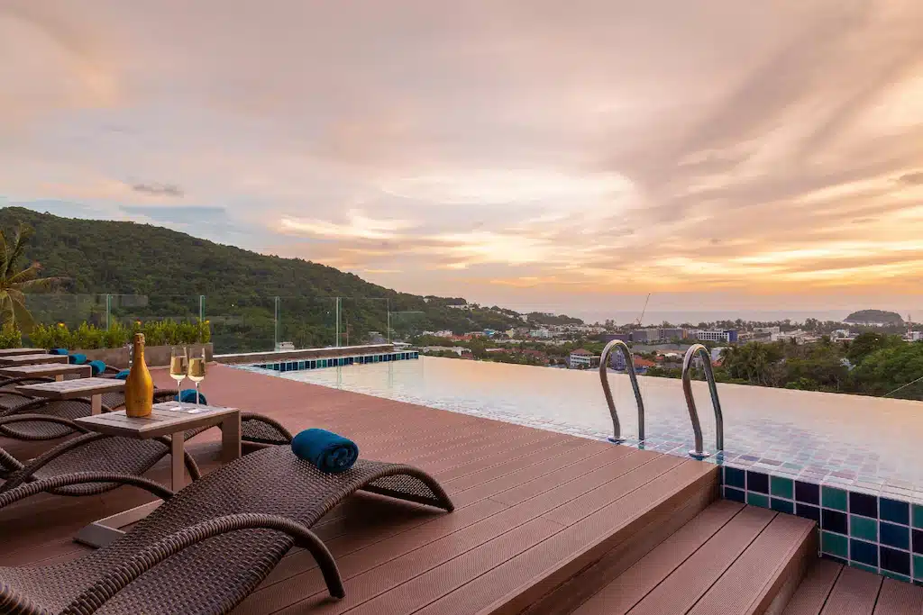 An infinity pool overlooks a scenic landscape at sunset, with a clear view of hills and the distant Big Buddha Phuket. Poolside, three lounge chairs with rolled blue towels and a small table holding a champagne bottle and two glasses add to the luxurious ambiance. The sky is painted with soft hues of orange and pink.