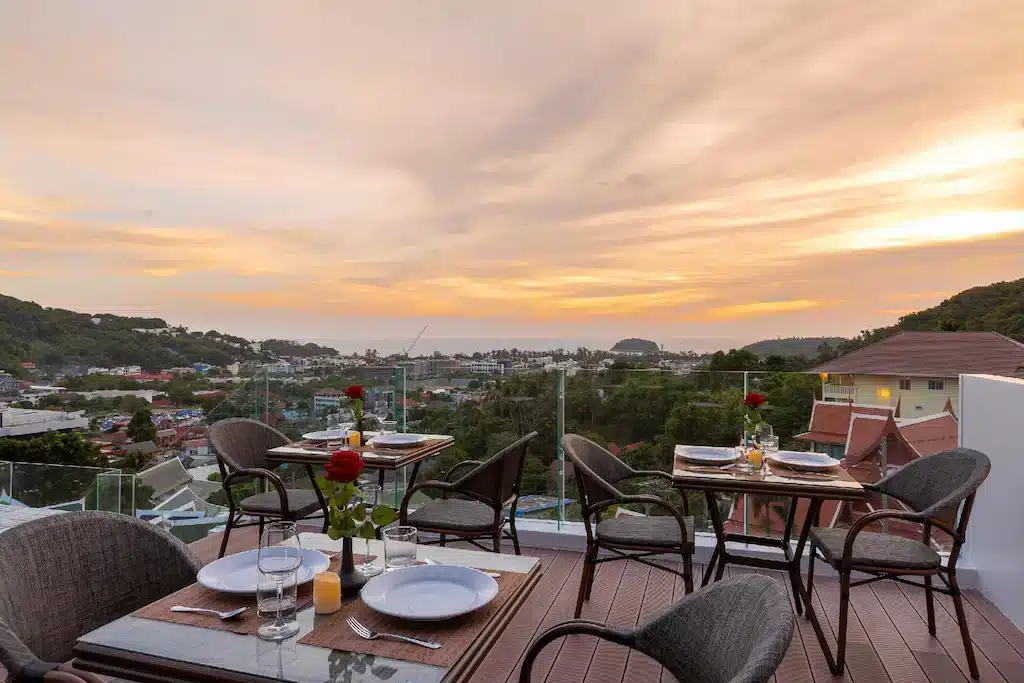 An outdoor rooftop dining area with several tables set for dinner, each adorned with a single red rose in a vase. The setting overlooks a scenic view of a town nestled in green hills, leading to a coastline, and you can even spot the majestic Big Buddha Phuket as the sun sets in shades of orange and pink.