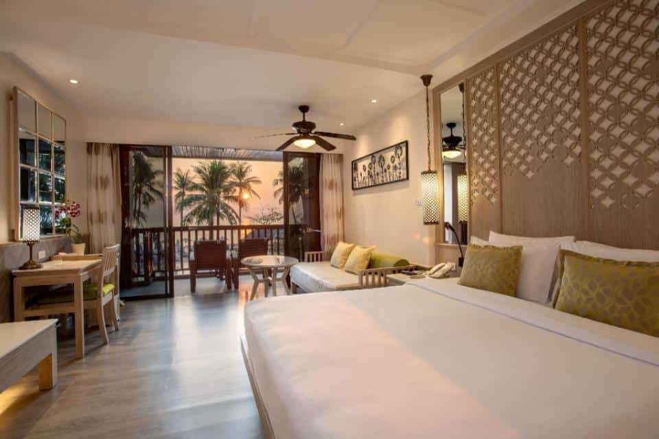 A spacious, well-lit hotel room features a large bed with white linens and green accent pillows. A seating area with a couch and coffee table sits by a sliding glass door that opens to a balcony with a view of palm trees and a sunset. Room decor includes modern furnishings and intricate lattice panels, perfect for guests seeking one of the best hotels in Phuket near the beach.