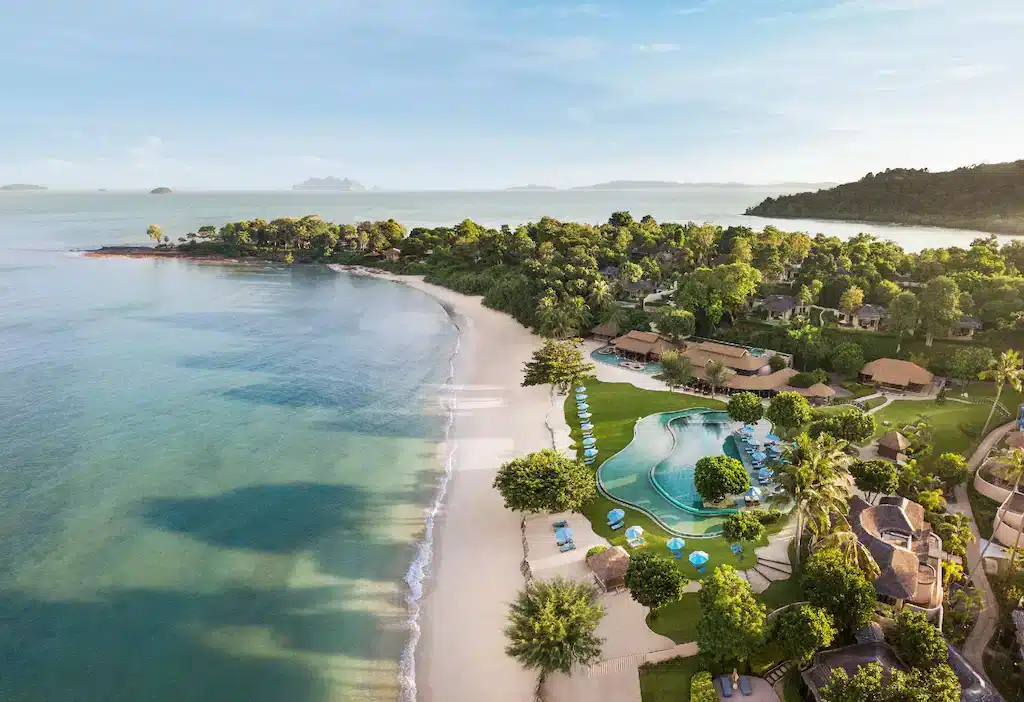 Aerial view of a tropical beach with clear blue water and white sand. A large resort, considered one of the best hotels in Phuket near the beach, occupies much of the coastline, featuring multiple pools, sun loungers, palm trees, and thatched-roof buildings. Dense greenery surrounds the resort. The horizon shows distant islands and a calm sea under a sunny sky.