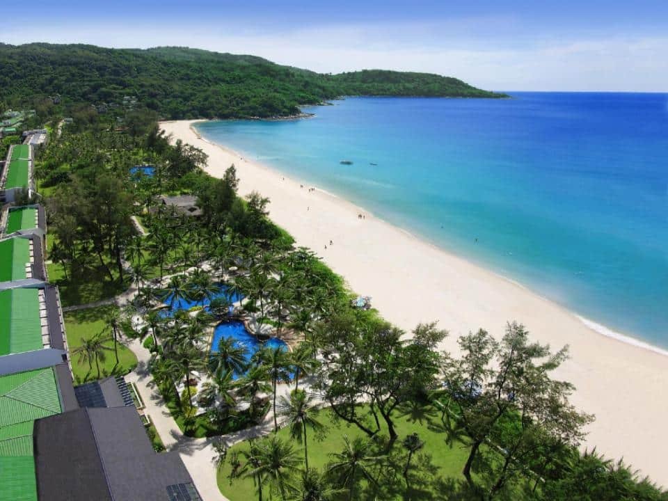 Aerial view of a coastal resort featuring a wide stretch of sandy beach with clear, turquoise waters. Lush green trees and tropical vegetation surround the luxury beachfront resort buildings and multiple swimming pools. The coastline extends into the distance, bordered by hills and dense forest.