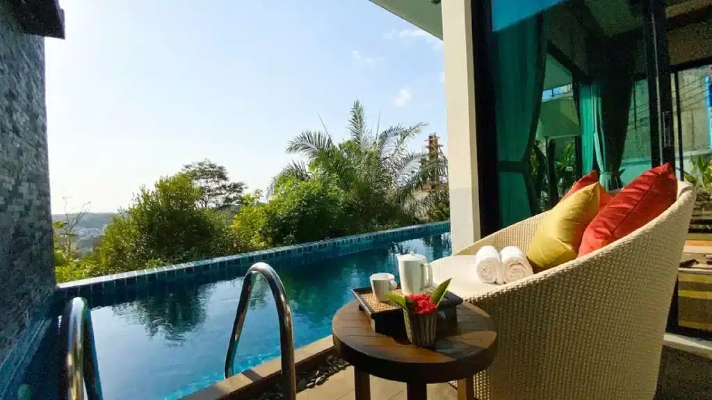 A luxurious outdoor scene featuring a pristine private swimming pool adjacent to a modern building with large glass windows. A wicker chair with colorful cushions and a small round table holding rolled towels, a teapot, and cups are placed poolside. Verdant foliage frames the view of Big Buddha Phuket under a clear sky in the background.