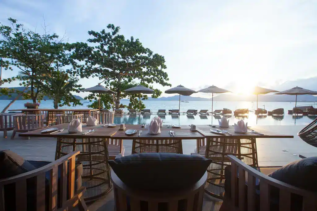 An outdoor dining area with wicker chairs and tables set with white napkins and cutlery overlooks an infinity pool at one of the best hotels in Phuket near the beach. In the background, there are sun loungers and umbrellas by the poolside. Beyond the pool, a body of water shimmers with a sunset behind trees on the horizon.