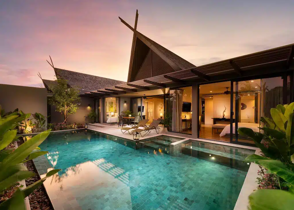 A luxurious villa with modern architectural design features large glass doors and a private pool. The interior is visible, showcasing an elegantly decorated bedroom and living area. The exterior has lush plants and trees. This villa belongs to one of the best hotels in Phuket near the beach, offering a serene sunset or sunrise backdrop.