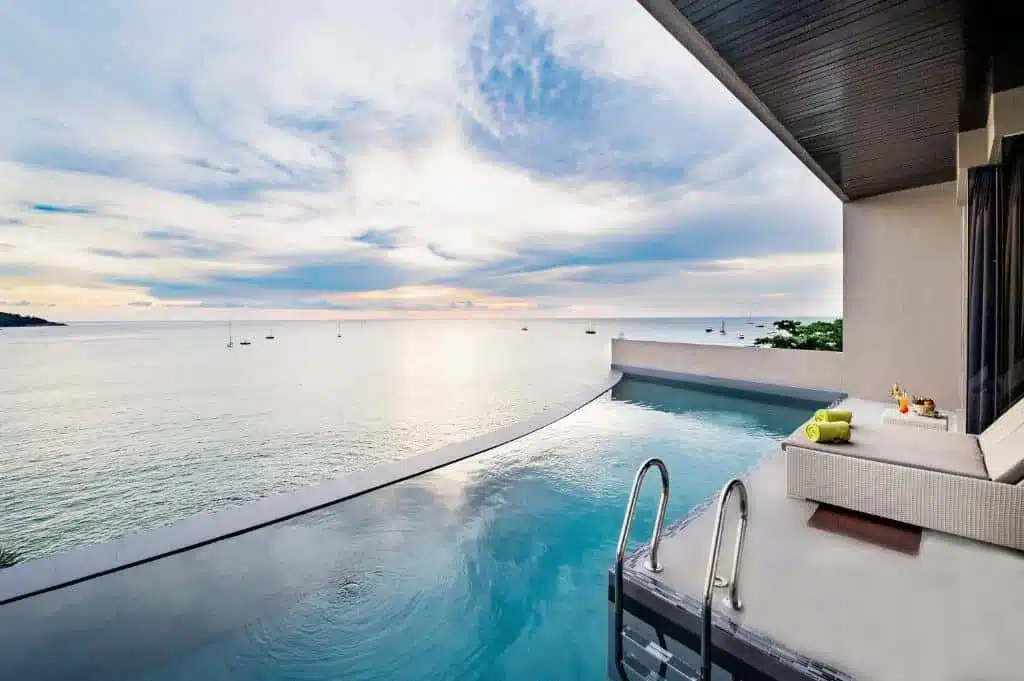 A modern infinity pool at one of the best hotels in Phuket near the beach overlooks a serene ocean with a few boats in the distance. The pool has metal steps leading into it, and a cushioned lounge area is nearby with rolled green towels and orange drinks. The partly cloudy sunset casts a peaceful ambiance.