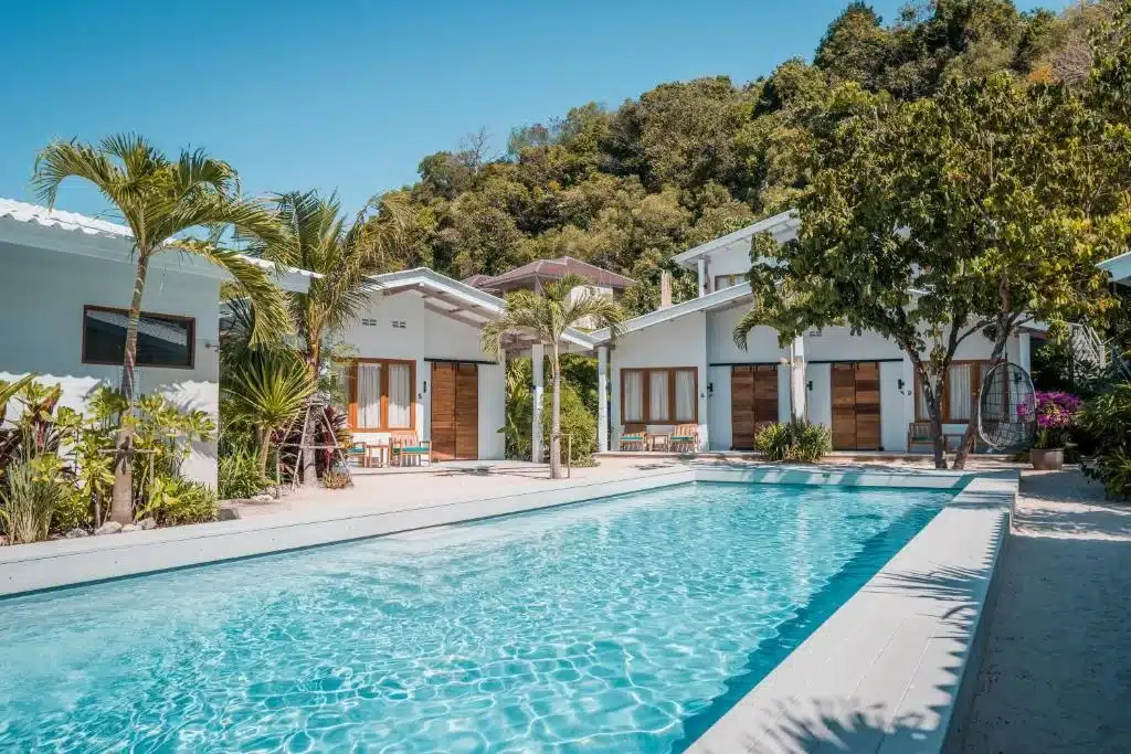 A serene outdoor scene features a clear blue swimming pool surrounded by white modern villas with wooden doors, characteristic of Phuket's beachfront resorts. The area is adorned with lush green palm trees and other tropical plants, creating a vibrant and inviting atmosphere. A backdrop of dense, green foliage-covered hills adds to the picturesque setting.