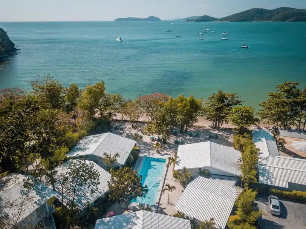 Aerial view of one of the luxury beachfront resorts in Phuket, showcasing white-roofed bungalows around a central outdoor pool. The resort is surrounded by lush greenery and overlooks a calm blue sea with several boats anchored in the distance. Rolling hills and additional islands are visible on the horizon under a clear sky.