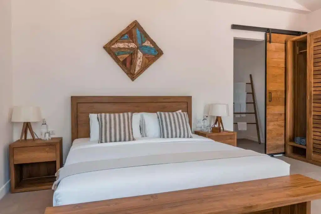 A cozy bedroom, reminiscent of the tranquil charm found in Phuket beachfront resorts, features a wooden bed frame with white and striped pillows and a light gray blanket. Wooden nightstands with lamps and water bottles flank the bed. Wall art hangs above the headboard, and a sliding wooden door reveals another room. A small wooden bench is at the bed's foot.