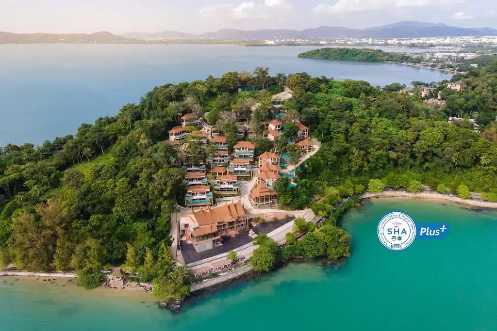 Aerial view of a luxury beachfront resort in Phuket, surrounded by lush green vegetation, situated on a small hill overlooking a calm, turquoise bay. The resort features several buildings with brown roofs. In the background, a larger body of water is visible with distant mountains and some urban structures. A "SHA Plus+" certification logo is placed near the lower right corner.