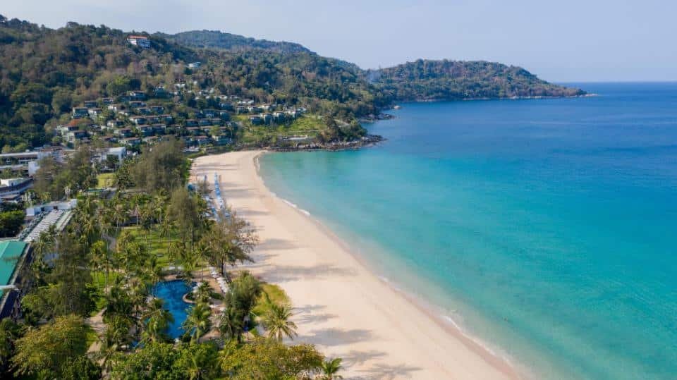 A pristine sandy beach with clear turquoise water is bordered by lush green hills and dotted with tropical palm trees. Modern buildings and luxury beachfront resorts in Phuket are nestled within the greenery on the hillside, and a large swimming pool is visible among the trees near the shoreline. The sky is clear.