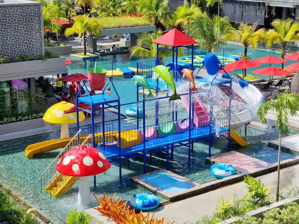 A colorful water playground in a luxury beachfront resort in Phuket features slides, a tipping bucket, and dolphin-shaped fountains. Surrounding the pool are lush green palm trees, sun loungers with red umbrellas, and other amenities. The lively pool area is both inviting and picturesque.