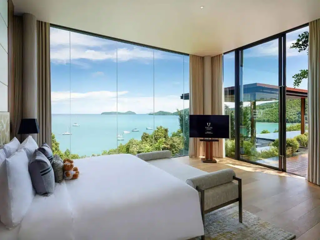 A luxurious bedroom at one of the best hotels in Phuket near the beach, featuring a large floor-to-ceiling glass window showcasing a stunning ocean view. The room boasts a neatly made king-size bed with white linens and plush pillows. A flat-screen TV is mounted on the wall, and the decor includes light wood and neutral tones.