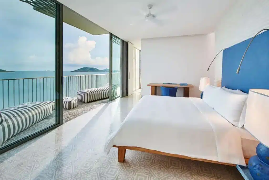 A modern, minimalist bedroom in one of the luxury beachfront resorts in Phuket features a large bed with white linens and a blue headboard. Floor-to-ceiling glass doors open to a balcony with striped lounge seats, offering a stunning ocean view. The room has a wooden desk, ceiling fan, and geometric-patterned floor tiles. The overall color scheme is blue and white.