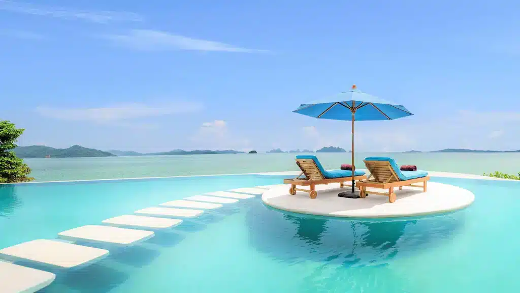 Infinity pool with stepping stones leading to a circular island featuring two wooden lounge chairs, blue cushions, and a matching blue umbrella. The pool overlooks a serene ocean with distant green islands under a clear blue sky. Lush greenery frames the pool on the left—an idyllic scene often found at the best hotels in Phuket near the beach.