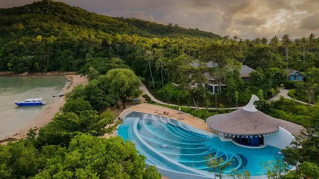 A serene tropical beach resort is among the best hotels in Phuket near the beach, featuring a large, uniquely designed pool with swirling patterns, enclosed by lush greenery. Nearby, a small sandy beach hosts a blue and white boat docked at the shore. A thatched-roof structure sits adjacent to the pool, backed by dense forested hills.