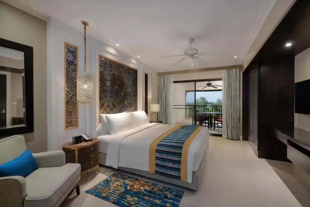 A modern hotel room featuring a cozy king-sized bed with white linens and a colorful striped runner. There is intricate artwork above the bed, a nightstand with an ornate lamp, a comfortable armchair with a blue pillow, and a rug with a floral pattern. As one of the best hotels in Phuket near the beach, it opens to a balcony with a view.