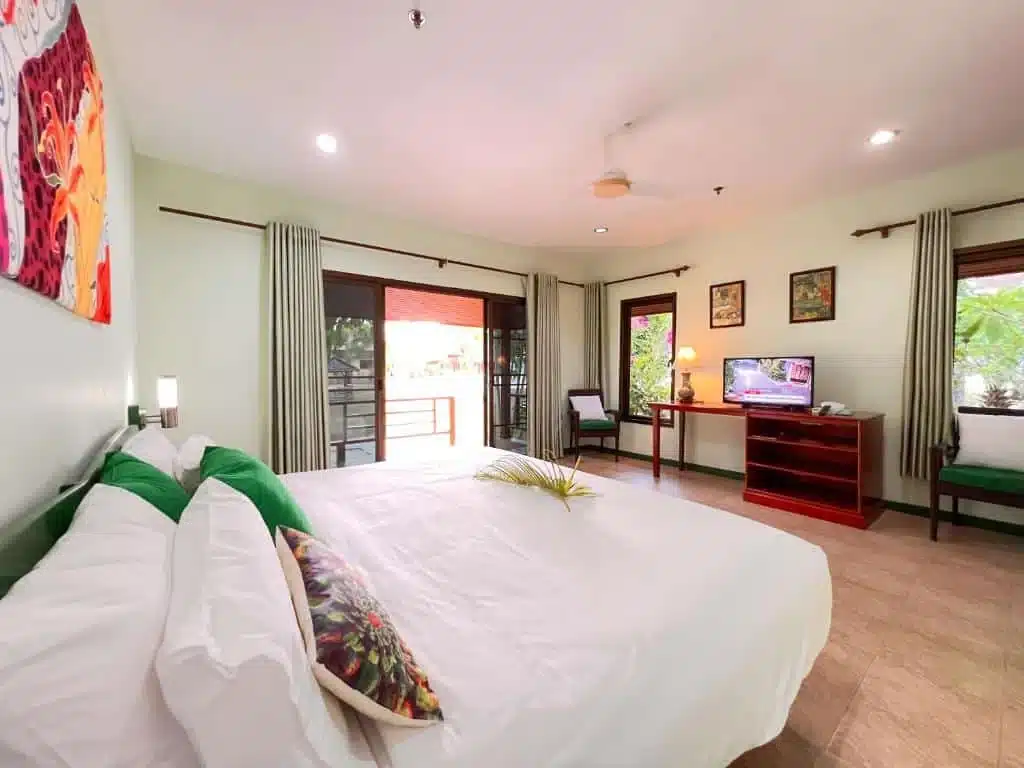 A spacious bedroom featuring a large bed with white linens and green floral pillows. There's a balcony with glass doors, beige curtains, a desk with a TV, and a chair. The walls are adorned with vibrant artwork and framed pictures. Staying here feels like being in one of the best hotels in Phuket near the beach.