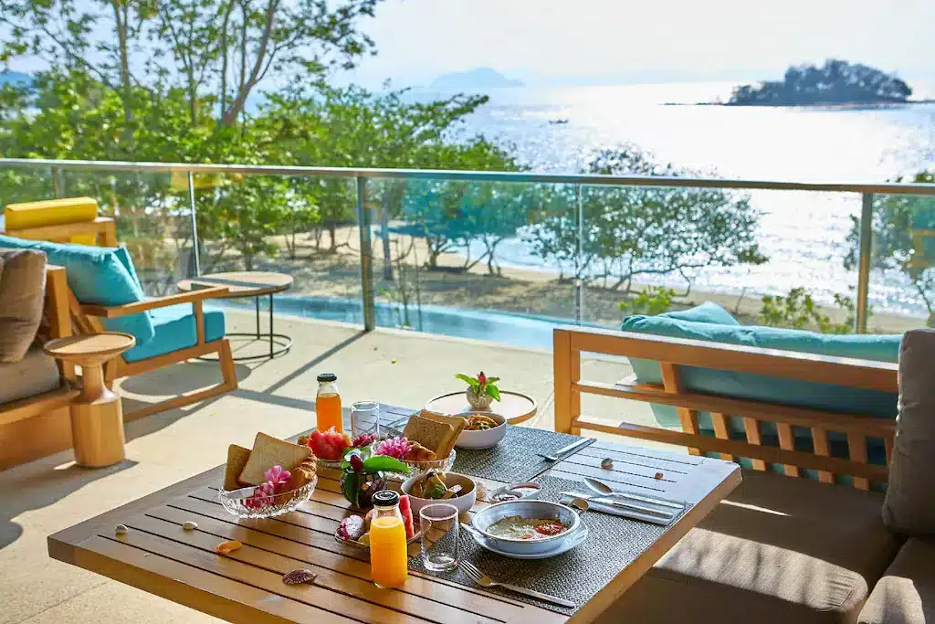 A balcony overlooking a scenic beach with a glass railing and patio seating at one of the best hotels in Phuket near the beach. The table is set with a meal, including bowls of food, bread, juice, water, and pink flowers. The background shows a sunlit ocean, islands, and lush trees. Cushioned chairs and a sofa with turquoise pillows are in the foreground.