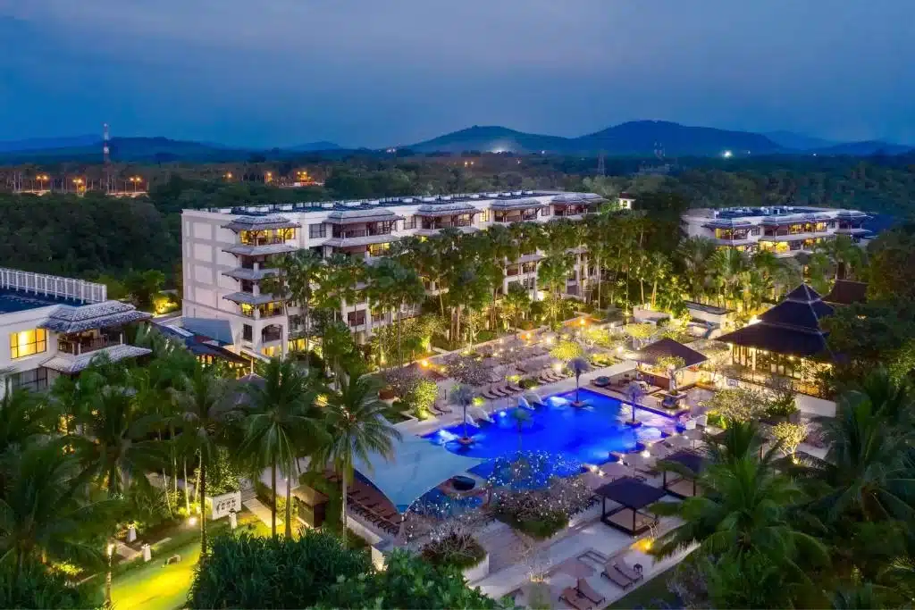 Aerial view of a luxury beachfront resort in Phuket at dusk. The image shows a large, multi-story building surrounded by tropical greenery. Centrally, there's a beautifully lit swimming pool area with lounge chairs and a small pavilion. Hills and a darkening sky serve as a serene backdrop in the distance.