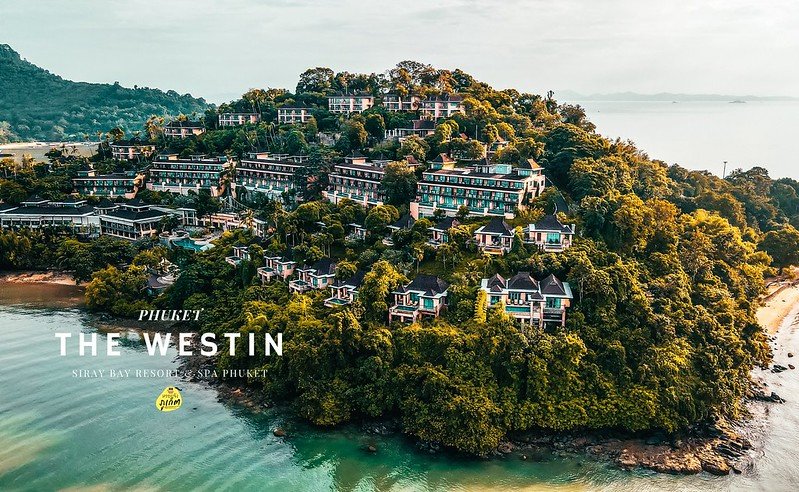 Aerial view of The Westin Siray Bay Resort & Spa in Phuket, one of the premier luxury beachfront resorts in Phuket, nestled on a lush hillside surrounded by blue waters. The resort features numerous buildings with white facades and turquoise roofs. Sandy beaches line the shoreline, while distant mountains are visible under a partly cloudy sky.