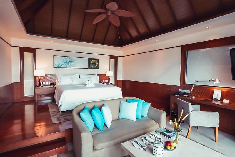 A luxurious hotel room, one of the best hotels in Phuket near the beach, features a king-size bed with white linens, flanked by nightstands with lamps. A beige sofa with blue and white pillows faces the bed. A desk and chair sit in the corner. Wooden-bladed ceiling fan hangs from the vaulted ceiling. Artwork adorns the walls, and a table holds fruits and decor.