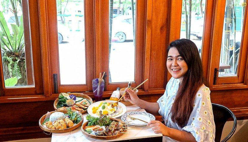 A smiling woman with long hair sits at a restaurant table laden with various dishes, including rice, vegetables, and soups. She holds chopsticks in her right hand and appears ready to eat. The background features large wooden windows with a view of greenery and Sarasin Bridge in the distance.