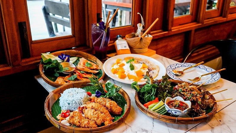 A table with various Thai dishes is set in a restaurant with wooden decor near the Sarasin Love Bridge. The dishes include fried rice with a purple flower garnish, assorted vegetables, salads, and fruit with sticky rice. Beverages are present, along with wooden utensils, and the setting appears cozy and inviting.