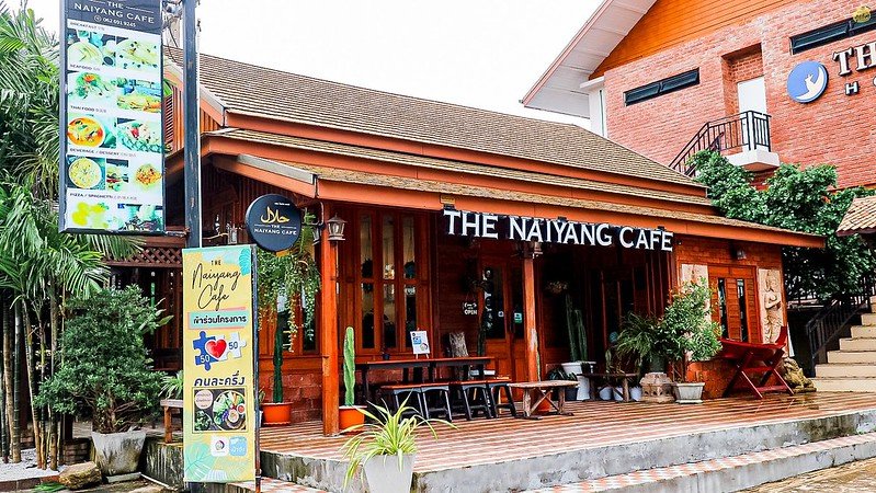 The image shows the exterior of The Naiyang Café, a wooden building with a sloped roof. There are large windows and a signboard displaying the café's name. To the left, a vertical menu board features colorful food pictures. Potted plants and outdoor seating adorn the entrance, evoking the charm of Sarasin Bridge nearby.