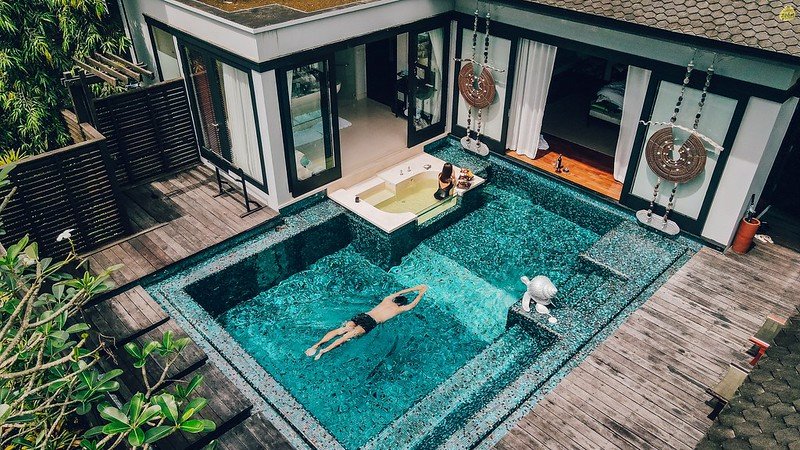 A luxurious villa features a private pool. A person swims in the large rectangular pool, while another sits in an elevated jacuzzi area. The villa, reminiscent of the elegance found near Sarasin Bridge, has an open-door design with wooden decking and greenery. Two hanging chairs are positioned near the sliding glass doors leading to the indoor space.