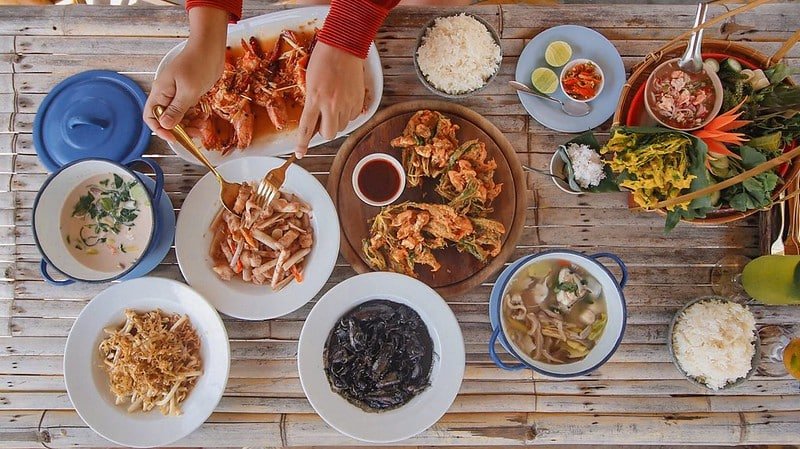 A person serving food at a table filled with various dishes, including seafood, rice, vegetables, and soups. The bamboo table is reminiscent of local charm near Sarasin Bridge. The dishes are arranged neatly with garnishes and condiments as the person's hands hold utensils, preparing to serve from a platter.