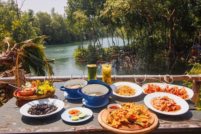 A scenic outdoor table is set with a variety of vibrant Thai dishes, including seafood, noodles, salads, and fried items. The backdrop features lush greenery and a serene river near the iconic Sarasin Love Bridge. Two tall drinks with straws are placed in the center, enhancing the tropical dining experience.