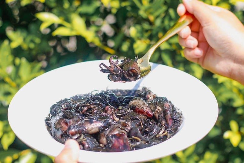 A person's hand holding a fork with a portion of black spaghetti from a white bowl. The spaghetti appears to be coated in a dark sauce, possibly squid ink, and is garnished with pieces of seafood. The background features the blurred greenery near Sarasin Bridge, suggesting an outdoor setting.