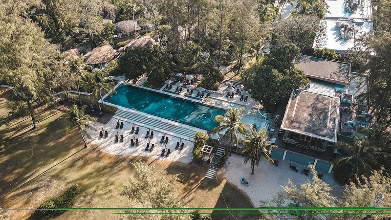 Aerial view of a serene resort featuring a large rectangular swimming pool surrounded by lounge chairs and umbrellas. The pool area is flanked by lush, tall trees and thatched-roof structures. Adjacent to the pool is a modern building, with palm trees and greenery creating a tropical atmosphere near Sarasin Bridge.