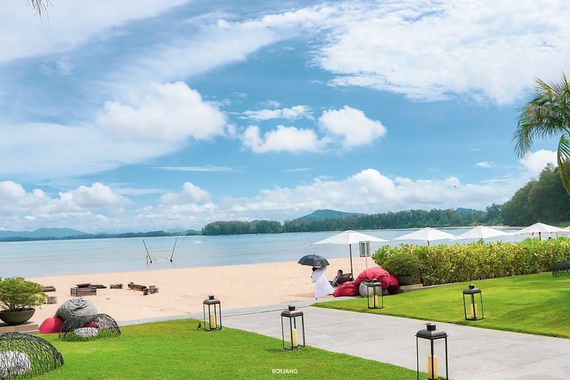 A serene beach scene with white sand, clear blue water, and a lightly clouded sky. A couple relaxes on red beanbag chairs under white umbrellas by the shore. In the foreground, a well-manicured grassy area with modern outdoor furniture and pathways is visible. The picturesque Sarasin Love Bridge extends in the distance, surrounded by lush greenery.