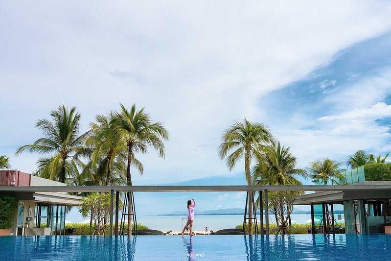 A person in a purple outfit walks across the Sarasin Bridge above a blue pool, surrounded by palm trees. Modern, glass-walled buildings are on either side, and a scenic ocean view with a partly cloudy sky forms the backdrop.