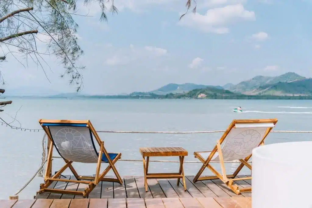 Two wooden lounge chairs with blue and white fabric seats face a calm, expansive lake on a wooden deck. A small wooden table sits between the chairs. Mountains and a partially cloudy sky are in the background. In the distance, reminiscent of Phuket beachfront resorts, a person rides a jet ski.