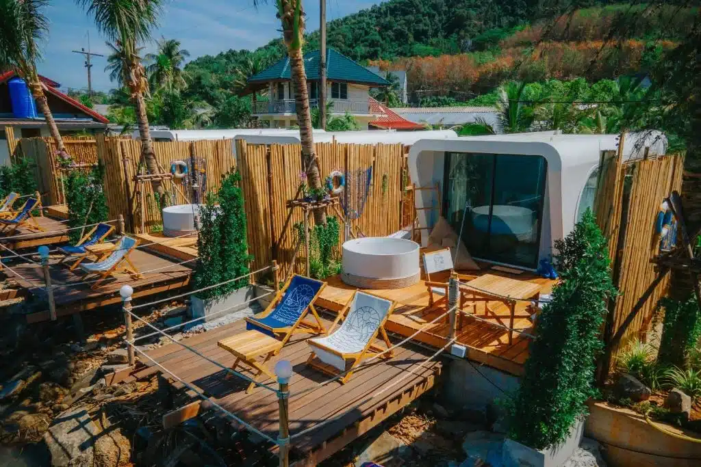 A cozy outdoor setting features modern, white cylindrical cabins with glass fronts. Wooden decks with blue and white lounge chairs, a circular hot tub, railings, and lush greenery provide scenic relaxation. This serene site rivals Phuket beachfront resorts with trees, a mountain background, and a sunny sky.