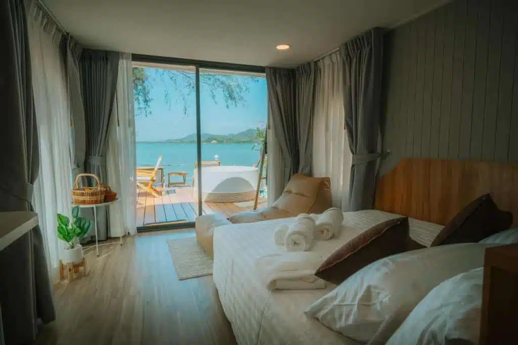 A cozy bedroom features a large bed with white linens and several pillows. Rolled towels rest at the foot of the bed. Grey curtains frame a glass sliding door that opens to a wooden deck overlooking a serene body of water. Outdoor seating and a wicker basket are visible on the deck, reminiscent of luxury beachfront resorts in Phuket.