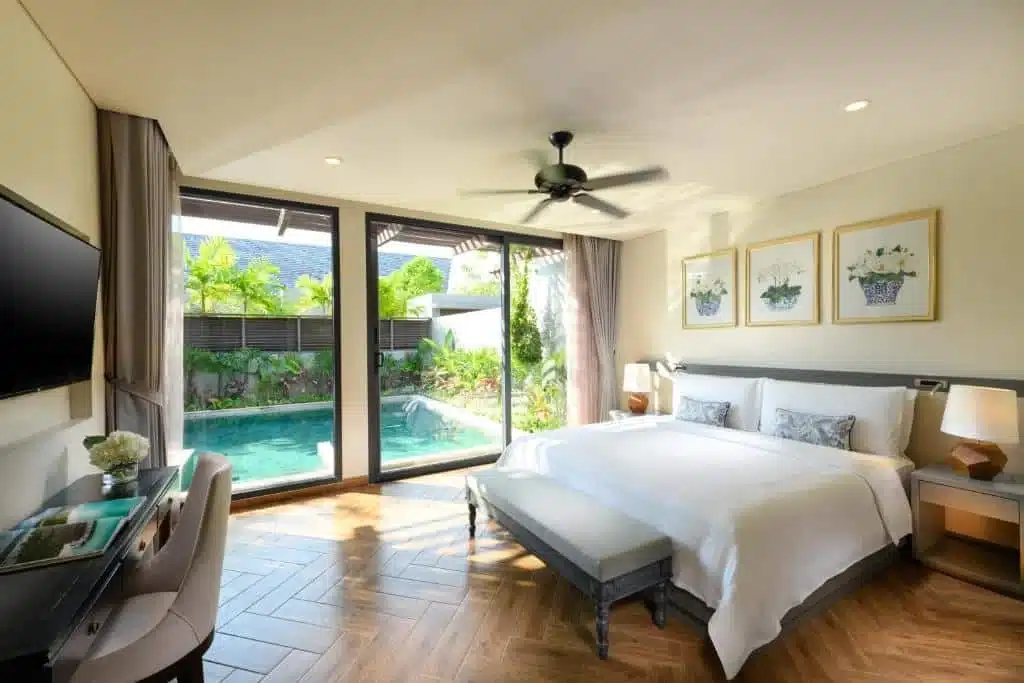 A spacious, bright bedroom with a large bed, white linens, and two side tables with lamps. The room features a flat-screen TV, a desk with a chair, and a ceiling fan. A glass sliding door leads to a private pool surrounded by greenery. Enjoy one of the best hotels in Phuket near the beach for your stay.