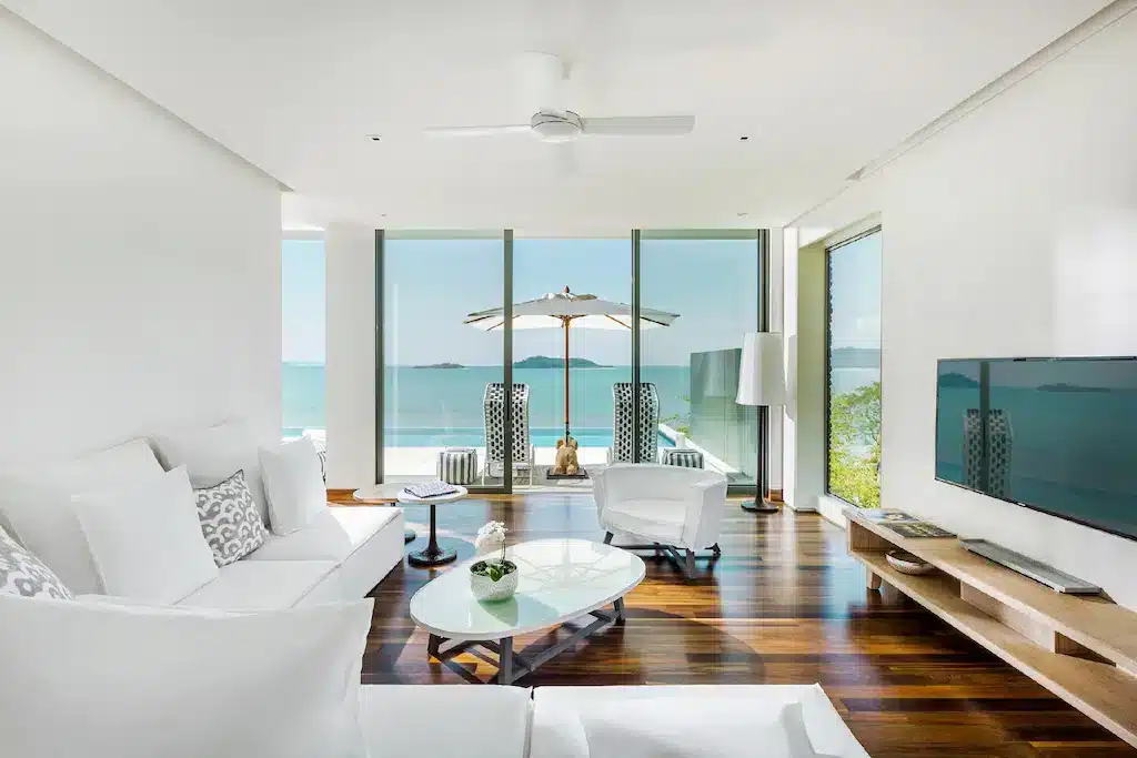 A modern, minimalist living room with white furniture, including a couch and armchairs. The space features large, floor-to-ceiling windows offering a stunning ocean view reminiscent of the best hotels in Phuket near the beach. A glass coffee table sits in the center, and a flat-screen TV is mounted on a wooden stand against the right wall.